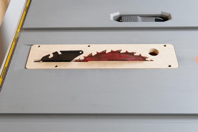 May include: A close-up of a table saw blade. The blade is red and has sharp teeth. It is set in a light brown insert. The table is gray and has a black handle.