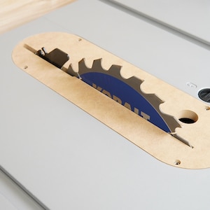 May include: A close-up of a table saw blade. The circular blade is partially visible, with a blue center and silver teeth. The blade is housed in a light brown, oval-shaped insert on a gray surface.