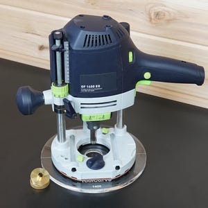 May include: A dark blue and light grey power router with lime green accents, featuring the text OF 1400 EQ on the side. The tool is mounted on a clear circular base plate labelled Toolcurve 1400, with a small brass circular guide bushing placed nearby.