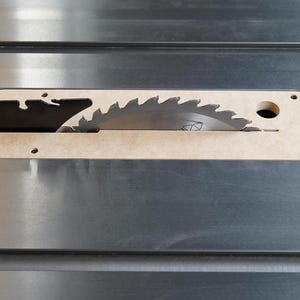 May include: Close-up of a table saw blade. The circular saw blade is partially visible through a beige insert in a silver metal surface. The blade has sharp teeth. The beige insert has a curved shape.
