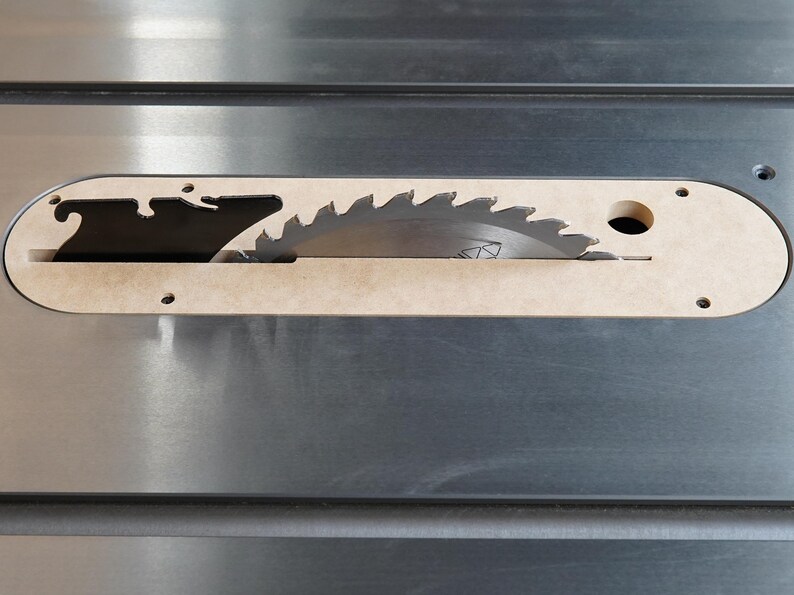 May include: Close-up of a table saw blade. The circular saw blade is partially visible through a beige insert in a silver metal surface. The blade has sharp teeth. The beige insert has a curved shape.