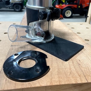 May include: A black and silver router with a clear plastic dust collection tube. The router is sitting on a wooden surface with a black base plate. There is a black plastic ring with a hole in the center.