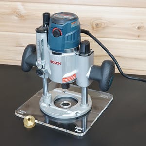 Puede incluir: Una fresadora de inmersión Bosch con carcasa de motor azul y plateada, dos mangos ergonómicos negros y una placa base de acrílico transparente. La herramienta incluye una escala de ajuste de profundidad, cable de alimentación y una pieza circular de latón.