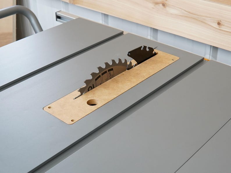 Puede incluir: Primer plano de una sierra de mesa con una hoja plateada y una inserci&oacute;n de madera. La hoja es parcialmente visible y est&aacute; rodeada por una superficie gris.