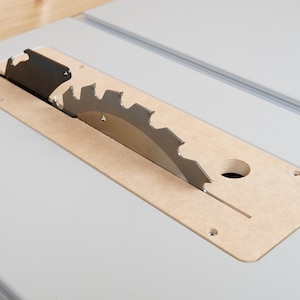 May include: Close-up of a table saw blade. The circular saw blade is silver and has sharp teeth. It is partially covered by a light brown insert with a circular hole and a slot. The saw is set on a light gray surface.
