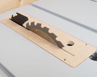 May include: Close-up of a table saw blade. The circular saw blade is silver and has sharp teeth. It is partially covered by a light brown insert with a circular hole and a slot. The saw is set on a light grey surface.