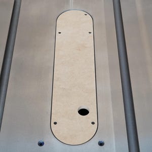 May include: A close-up shot of a light-colored wooden insert, set into a silver-colored metal surface. The insert is oval-shaped with a circular hole and several smaller holes. The metal surface has two parallel grooves.