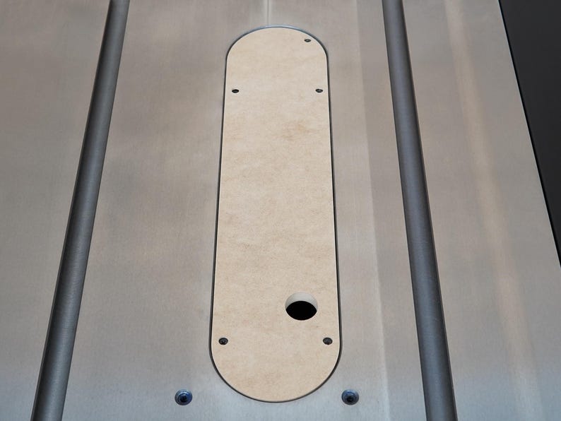 May include: A close-up shot of a light-colored wooden insert, set into a silver-colored metal surface. The insert is oval-shaped with a circular hole and several smaller holes. The metal surface has two parallel grooves.