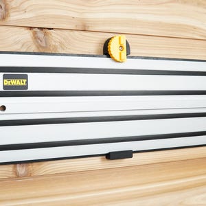 Op de afbeelding: Een witte DEWALT geleiderail voor een railsäge met zwarte strepen en een gele knop. De rail is gemonteerd aan een houten muur.