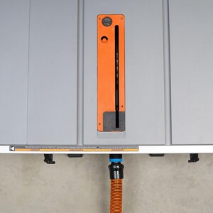 Op de afbeelding: Een grijze tafelzaag met een oranje stofafvoer en een zwart-blauwe stofafzuigbuis.
