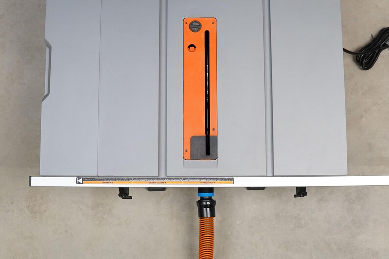 Op de afbeelding: Een grijze tafelzaag met een oranje stofafvoer en een zwart-blauwe stofafzuigbuis.