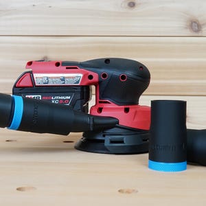 Puede incluir: Una lijadora inalámbrica roja y negra con una manguera negra y dos accesorios negros con detalles azules. La lijadora tiene una batería que dice "REDLITHIUM XC5.0". Las herramientas están sobre una superficie de madera marrón claro.