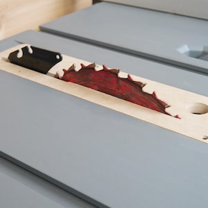 May include: Close-up of a table saw blade. The circular saw blade is red and has sharp teeth. The blade is set in a light-colored wooden insert, which is set in a gray table.