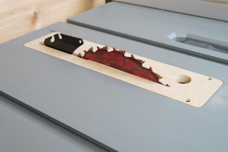 May include: Close-up of a table saw blade. The circular saw blade is red and has sharp teeth. The blade is set in a light-colored wooden insert, which is set in a gray table.