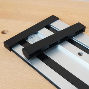 May include: A metal and black woodworking track system. The system features a silver track with black accents and two black sliding components. The track is set on a light wood surface.