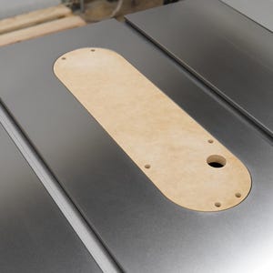 May include: A close-up shot of a table saw with a light brown, oval-shaped insert. The insert has several small holes and a larger circular opening. The saw's surface is a metallic gray color.