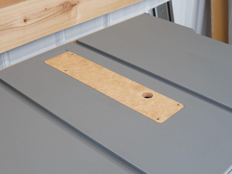 Puede incluir: Primer plano de una sierra de mesa gris con un inserto de madera. El inserto tiene un agujero en el centro y est&aacute; rodeado de una superficie gris.