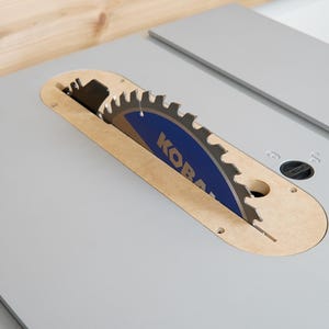 May include: A table saw with a circular blade. The blade is blue and silver with the brand name "KOBALT" printed on it. The saw is set in a light gray surface with a wooden blade guard.