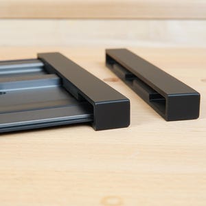 May include: A close-up shot of a black metal track system with two rectangular components. The system is designed for woodworking or similar applications. The components are positioned on a light-colored wooden surface.