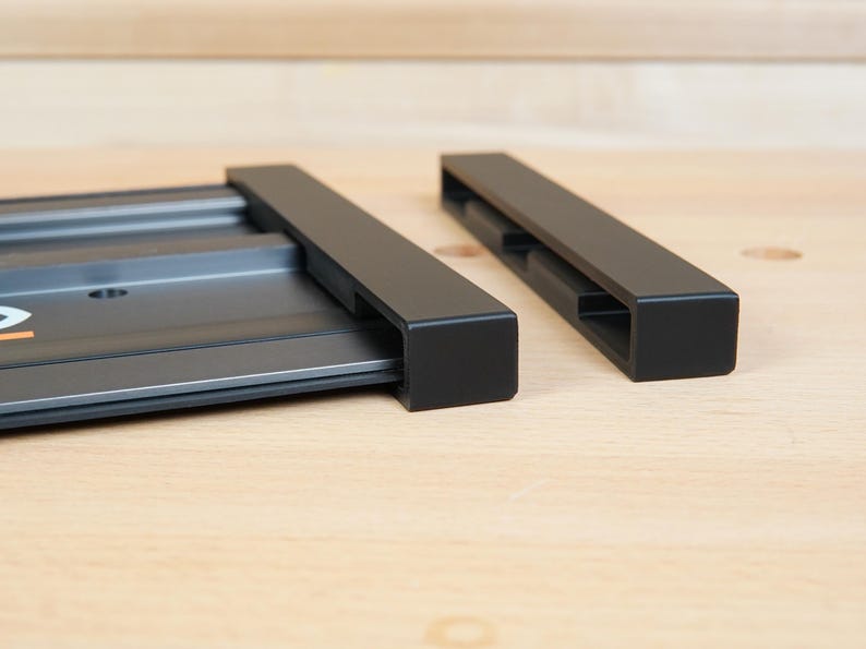 May include: A close-up shot of a black metal track system with two rectangular components. The system is designed for woodworking or similar applications. The components are positioned on a light-colored wooden surface.