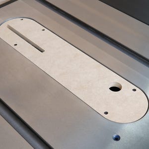 May include: A close-up view of a table saw with a light-colored insert. The insert is oval-shaped with a slot and a hole. The saw's surface is metallic gray, and the insert is secured with screws.
