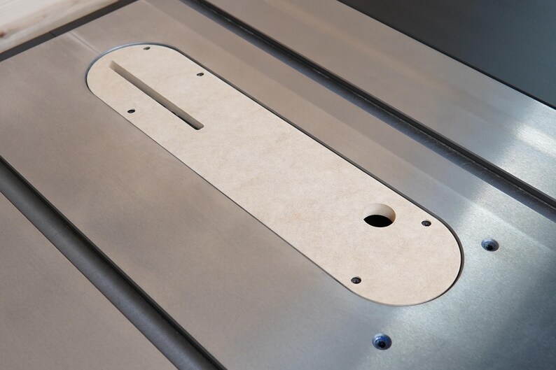 May include: A close-up view of a table saw with a light-colored insert. The insert is oval-shaped with a slot and a hole. The saw's surface is metallic gray, and the insert is secured with screws.