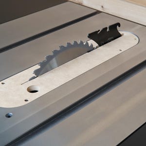 May include: Close-up of a table saw with a silver blade and a black blade guard. The saw blade is circular with sharp teeth. The table is gray with a light-colored insert around the blade.