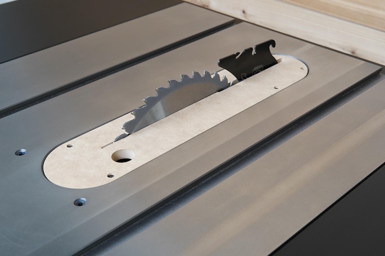 May include: Close-up of a table saw with a silver blade and a black blade guard. The saw blade is circular with sharp teeth. The table is gray with a light-colored insert around the blade.