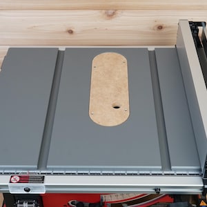 Może przedstawiać: Szara piła stołowa z szarym blatem i srebrną prowadnicą. Blat ma okrągły otwór na tarczę i prostokątny otwór na prowadnicę kątową. Prowadnica jest regulowana i ma skalę do pomiaru.