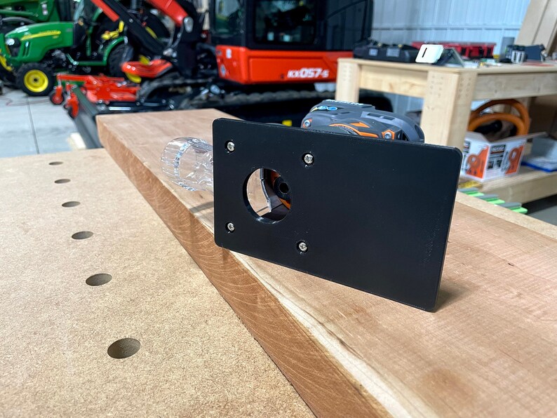 Puede incluir: Una plantilla de router negra con un recorte circular para una fresa. La plantilla est&aacute; hecha de pl&aacute;stico y tiene cuatro tornillos que la sujetan. La plantilla est&aacute; sobre una mesa de trabajo de madera.