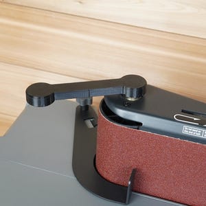 May include: A black belt sander with a red abrasive belt. The sander has a black handle and a gray base. The image is taken from a high angle, showing the details of the tool.