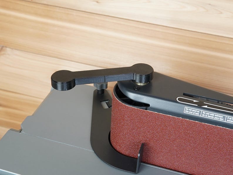 May include: A black belt sander with a red abrasive belt. The sander has a black handle and a gray base. The image is taken from a high angle, showing the details of the tool.