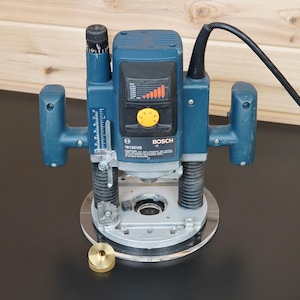 Könnte beinhalten: Eine blaue Bosch 1613EVS Oberfräse mit zwei schwarzen Griffen, einer Tiefeneinstellskala und einem gelben Drehzahlregler. Das Elektrowerkzeug verfügt über eine kreisförmige Grundplatte und ein kleines messingfarbenes Führungsteil davor.