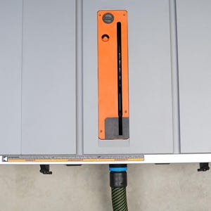 Puede incluir: Una sierra de mesa gris con un puerto de recolecci&oacute;n de polvo naranja y un cable de alimentaci&oacute;n negro. Una manguera de recolecci&oacute;n de polvo verde y negra est&aacute; conectada al puerto.