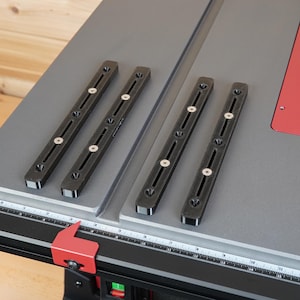 May include: A gray table saw with black rails and a red accent. The saw features a ruler along the front edge and is set against a wooden background. The saw's components are arranged on a gray surface.
