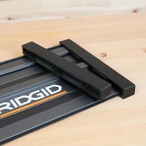May include: A black and gray RIDGID track saw guide rail system. The system includes a black metal base with the RIDGID logo in white and orange, and two black guide rails. The system is designed for woodworking.