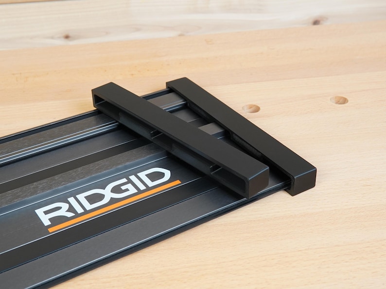 May include: A black and gray RIDGID track saw guide rail system. The system includes a black metal base with the RIDGID logo in white and orange, and two black guide rails. The system is designed for woodworking.