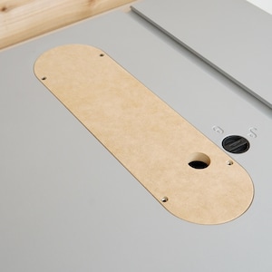 May include: A light gray table saw with a natural wood-colored insert. The insert has a rounded end and a hole. A black knob and a gray ruler are also visible.