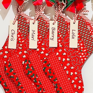 May include: Four red and white Christmas stockings with a snowman and candy cane pattern. Each stocking has a personalised wooden tag with the names Chris, Mari, Barry, and Lola.
