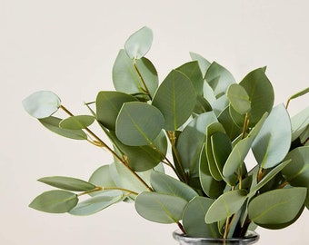 Op de afbeelding: Een bos kunstmatige eucalyptusbladeren in een transparante glazen vaas. De bladeren zijn lichtgroen en hebben een realistisch uiterlijk.