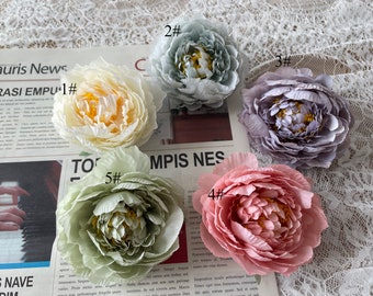 Puede incluir: Cinco cabezas de flores de peonía artificiales en varios colores, incluyendo blanco, verde claro, gris, rosa y un verde más oscuro. Las flores están dispuestas sobre una superficie de encaje blanco y un fondo de periódico. Las flores están numeradas del 1 al 5.