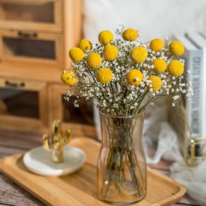Op de afbeelding: Een boeket gedroogde gele billy buttons in een heldere glazen vaas op een houten dienblad. De bloemen zijn gerangschikt met witte baby's breath.