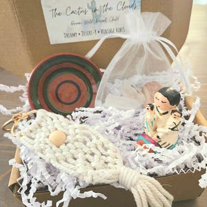 May include: A gift box containing a macrame keychain, a small ceramic plate with red and green stripes, a small figurine, and a sheer bag. The box has a card that reads "A Dreamy Little Gift Just For You... The Cactus in the Clouds."