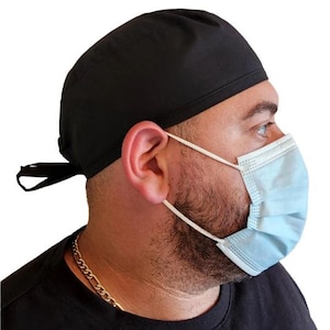 Op de afbeelding: Een zwarte scrubcap met een stropdas, gedragen door een persoon met een baard en een blauw mondkapje.