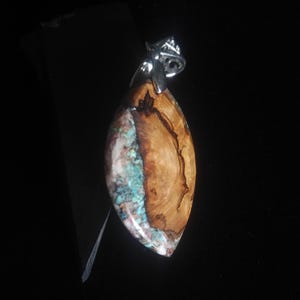 May include: A handcrafted pendant with a unique design. The pendant features a polished, teardrop-shaped wooden section with natural grain patterns, paired with a section of turquoise-colored stone. A silver-colored bail is attached at the top.
