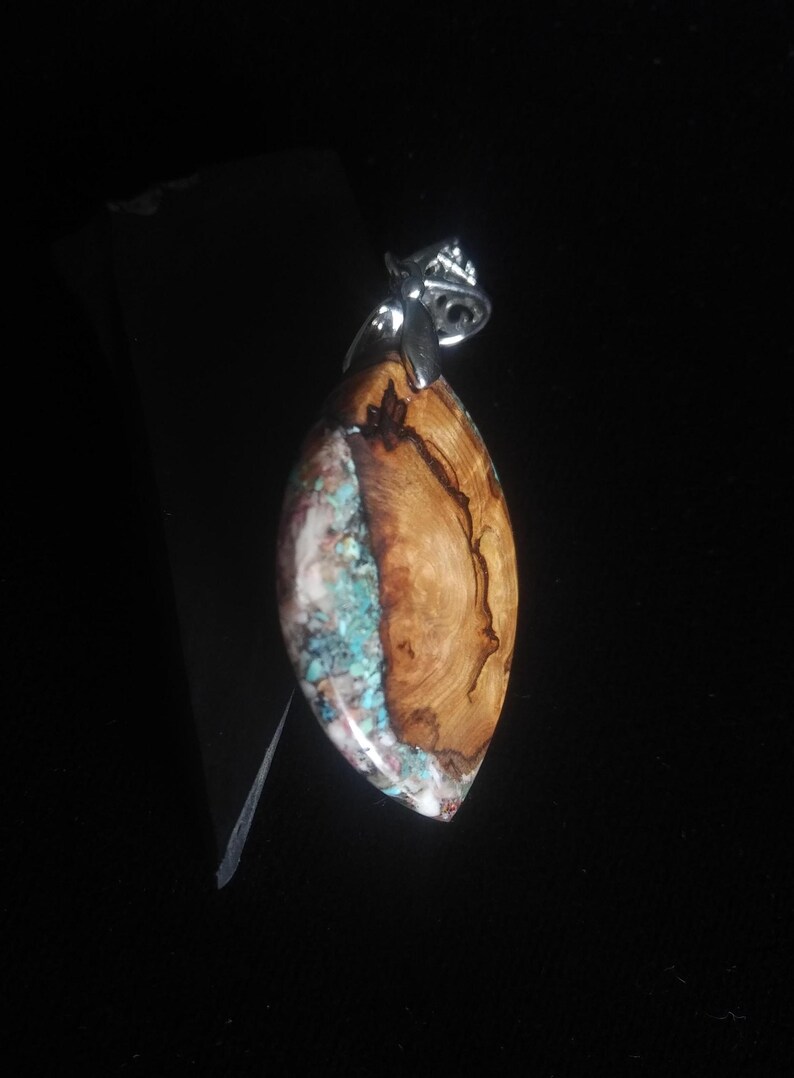 May include: A handcrafted pendant with a unique design. The pendant features a polished, teardrop-shaped wooden section with natural grain patterns, paired with a section of turquoise-colored stone. A silver-colored bail is attached at the top.