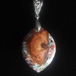 May include: A handcrafted pendant with a silver bail. The focal point is a marquise-shaped stone, featuring a wood grain center surrounded by turquoise, red, and white accents. The pendant is suspended against a black background.