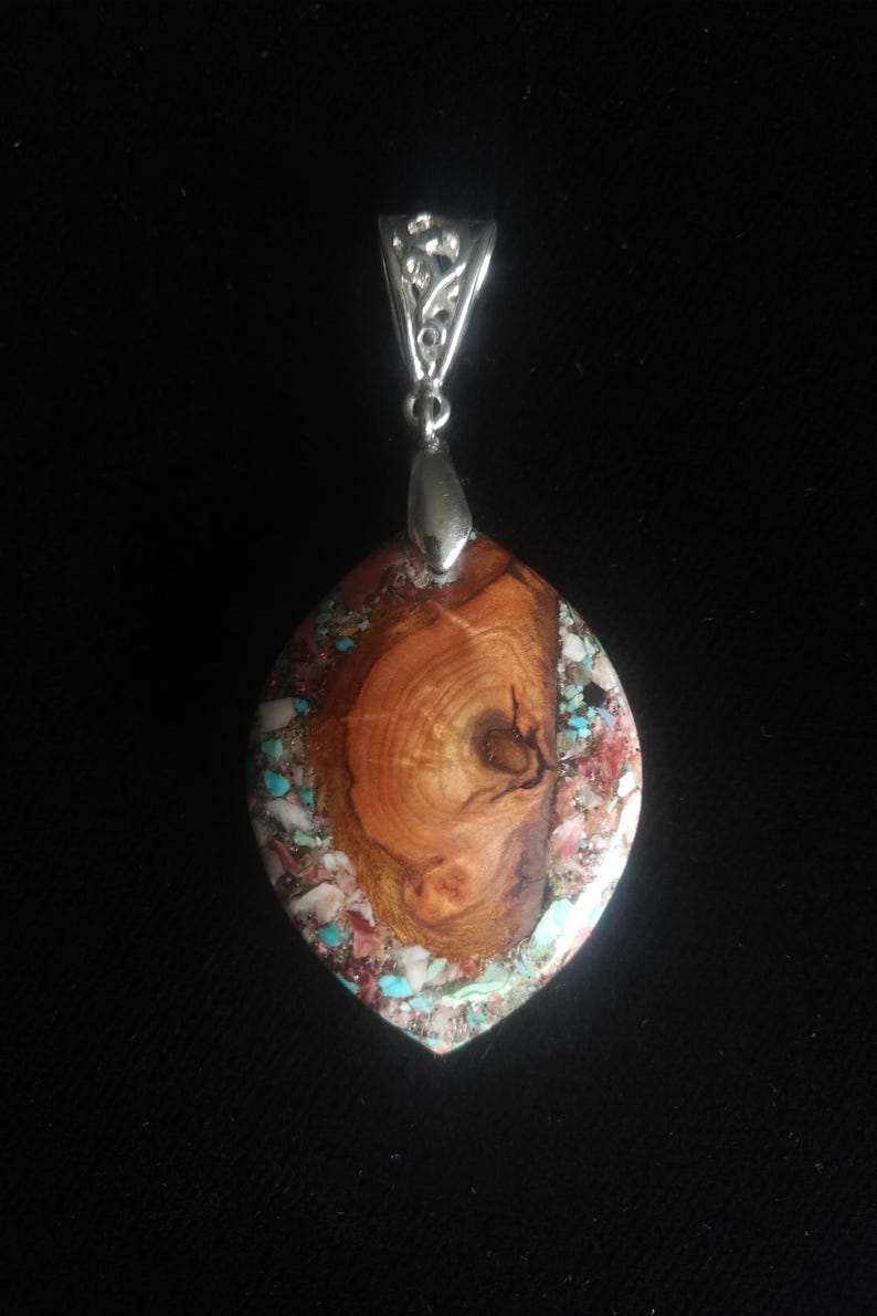 May include: A handcrafted pendant with a silver bail. The focal point is a marquise-shaped stone, featuring a wood grain center surrounded by turquoise, red, and white accents. The pendant is suspended against a black background.