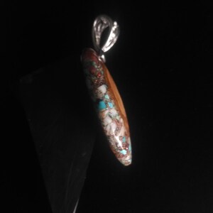 May include: A pendant with a silver bail and a teardrop-shaped stone. The stone features a mosaic pattern of brown, white, turquoise, and red-orange colors. The pendant is likely a piece of jewelry.