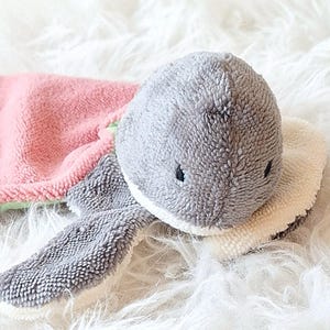 Könnte beinhalten: Ein plüschiger, grau geformter Baby-Waschlappen in Form eines Hais mit einem rosa und grünen Handtuchkörper. Der Hai hat kleine schwarze Augen und einen weißen, flauschigen Kragen. Der Waschlappen ist für Babys weich und sanft.
